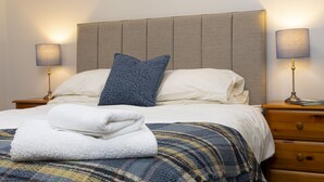 Una cama bien tendida con una almohada azul, sábanas blancas y una toalla doblada. Hay dos mesitas de noche con lámparas.