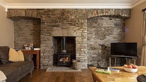 A cozy living room with a stone fireplace, a comfortable sofa, a wooden coffee table with books and a bowl of fruit, and a flat-screen TV. - Yr Hen Ysgol - historic cottage in grounds of manor house (Swansea)