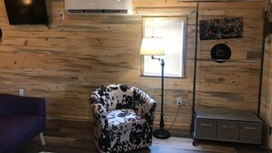 A room with wooden walls, a mounted TV, a ceiling fan, a window with blinds, a patterned armchair, a floor lamp, and a small cabinet. - Cabin #9 | 10 Min to Glacier Park Entrance, Minutes to Glacier Park! (Coram)