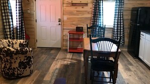 A cozy cabin interior with a wooden interior, a dining table with chairs, a sofa, a kitchen area with a refrigerator, and plaid curtains. - Cabin #9 | 10 Min to Glacier Park Entrance, Minutes to Glacier Park! (Coram)