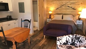 A compact room with a wooden floor, a bed with a headboard, a dining table with chairs, a small kitchenette, and a nightstand with a lamp. - Cabin #9 | 10 Min to Glacier Park Entrance, Minutes to Glacier Park! (Coram)