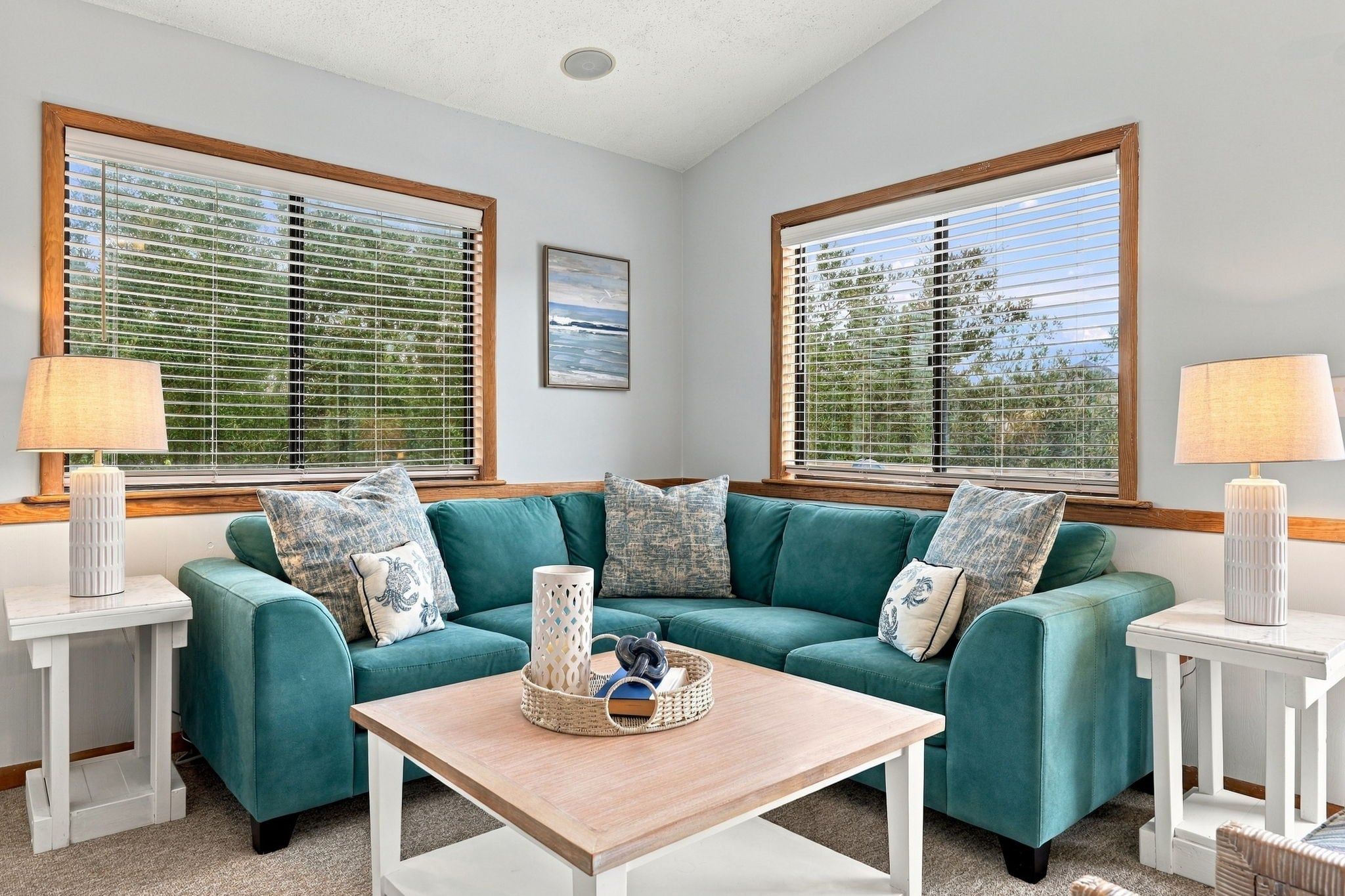A living room with a teal sofa, two side tables, a coffee table, and large windows with blinds.