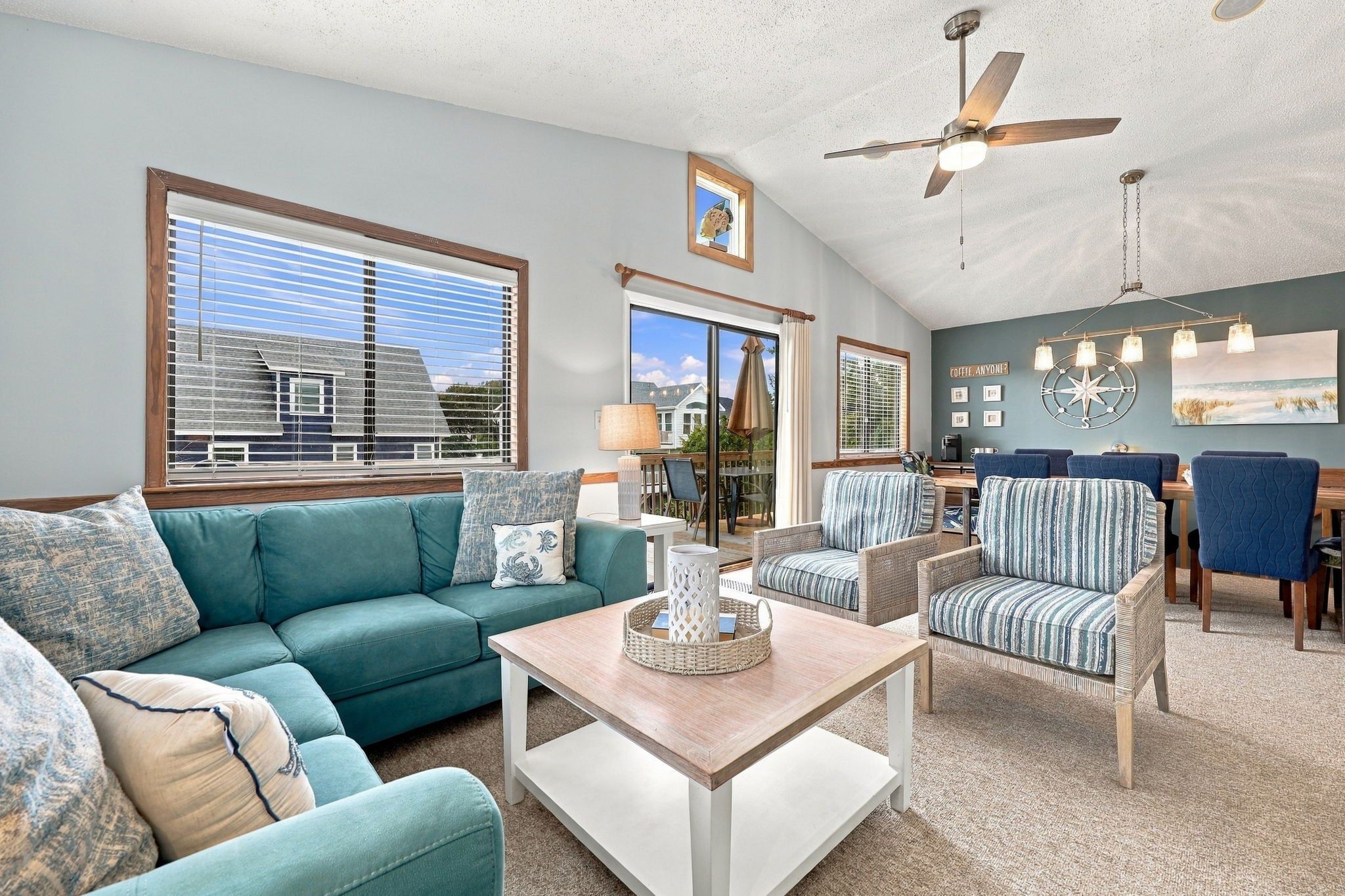 A spacious living room with a large window, a ceiling fan, and a dining area with blue upholstered chairs.