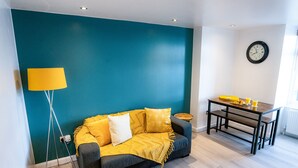 A modern living room with a teal accent wall, a sofa with yellow cushions, a floor lamp, and a dining table with a yellow bowl. - Perfect flat for the beach and town centre (Bournemouth)