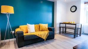 A modern living room with a teal accent wall, a dark grey sofa with yellow and white cushions, a yellow floor lamp, and a small table with drinks. - Perfect flat for the beach and town centre (Bournemouth)