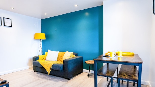 A modern living room with a blue accent wall, a grey sofa with yellow cushions, a wooden coffee table, and a small round table with a lamp. - Perfect flat for the beach and town centre (Bournemouth)