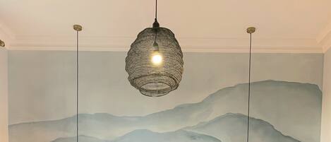 A bedroom with a large bed, two bedside tables with lamps, a hanging light fixture, and a mountain-themed wall mural.
