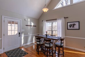A dining area with a wooden table and four chairs, a chandelier, and a window with curtains.