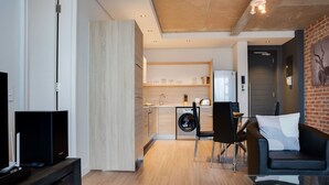 A modern living room with a black sofa, a glass coffee table, and a TV on a wooden stand. A kitchen is visible in the background with a washing machine and a dining area. - Modern 1 Bedroom Apartment in Century City (Cape Town)