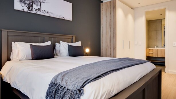 A modern bedroom with a large bed, a bedside table with a lamp, and a view into the bathroom. - Modern 1 Bedroom Apartment in Century City (Cape Town)