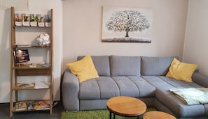 A living room with a grey sectional sofa, two yellow pillows, a small wooden coffee table, and a bookshelf with various items.