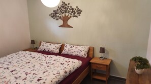 A bedroom with a bed, two nightstands, a lamp on each, a wooden tree wall decoration, and a potted plant.