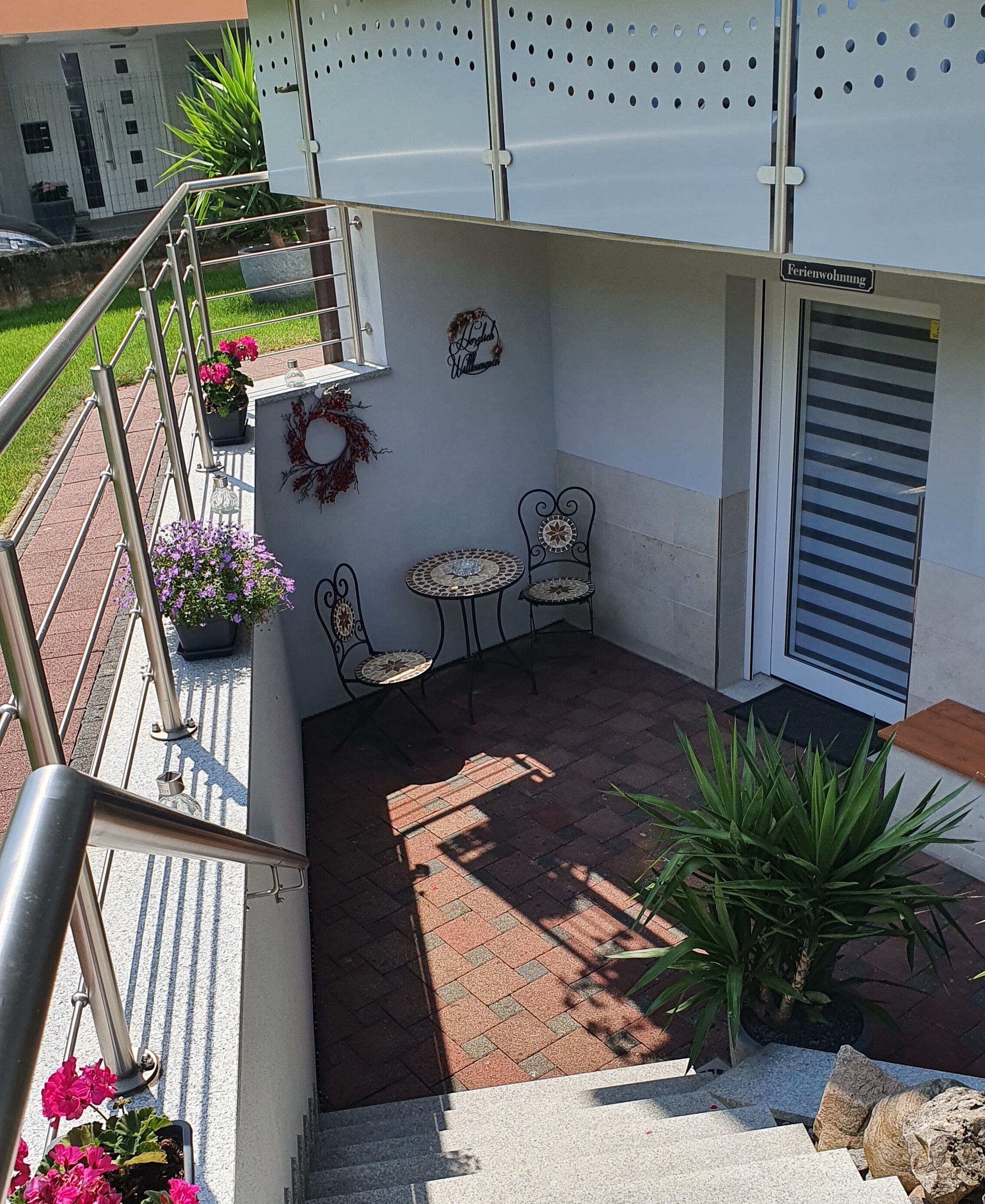 A small outdoor patio area with a metal railing, two metal chairs, a small round table, and potted plants.