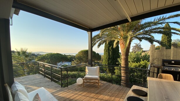 A covered outdoor deck with a wooden floor, a white sofa with cushions, a wooden table with black cushions, and a grill. - Golfe de Saint-tropez, Villa Contemporaine Avec Piscine, vue mer et Plage à Pied (Cavalaire-sur-Mer)