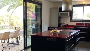 A modern kitchen with a central island, a dining area with a table and chairs, and a sliding glass door leading to an outdoor patio. - Golfe de Saint-tropez, Villa Contemporaine Avec Piscine, vue mer et Plage à Pied (Cavalaire-sur-Mer)