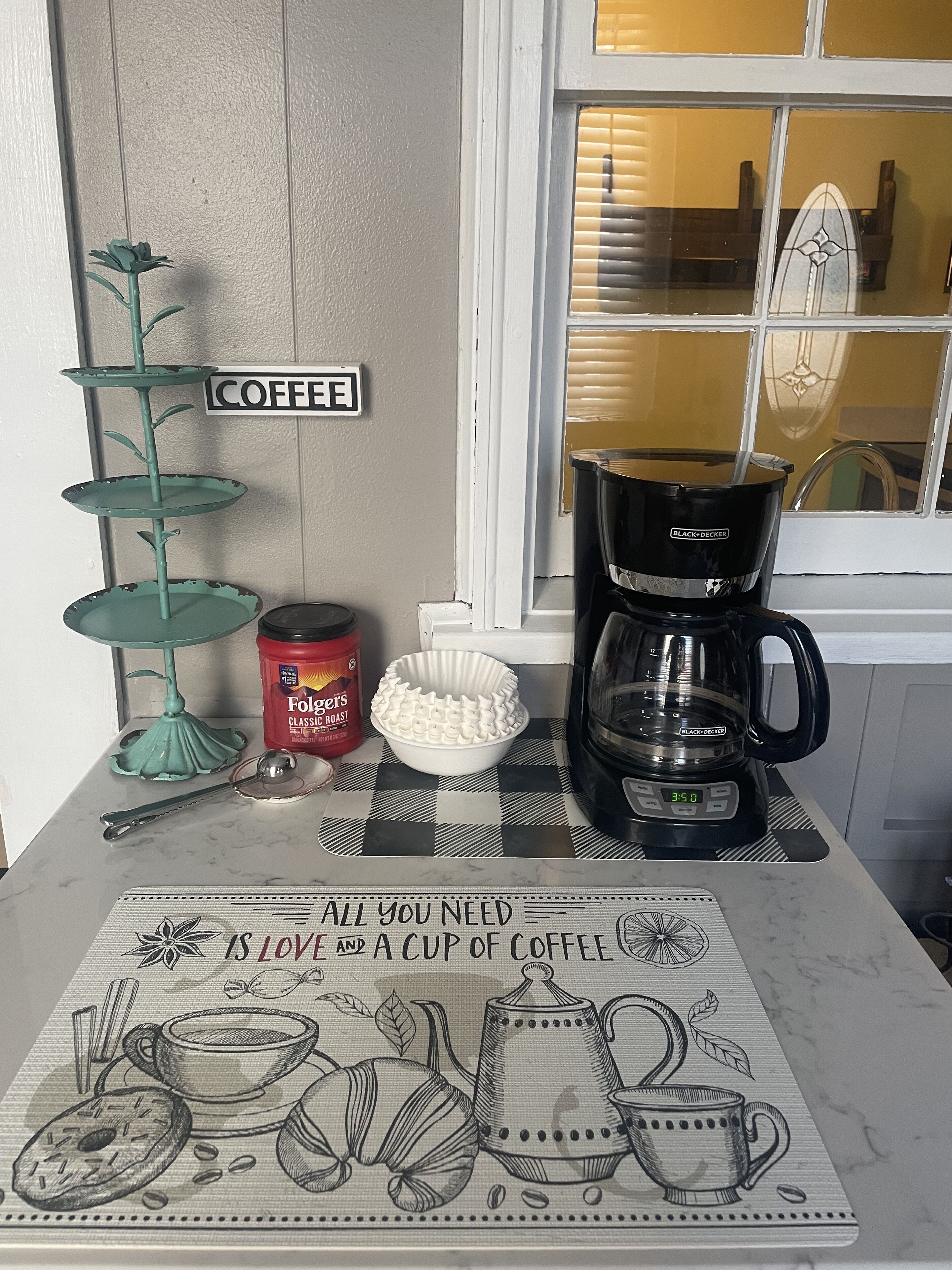 Una encimera de cocina con una cafetera, café Folgers Classic Roast, un molde para muffins y un cartel decorativo que dice "COFFEE".