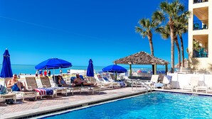 Un área junto a la piscina con sillones de descanso y sombrillas, piscina y palmeras.
