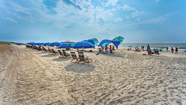 A beach with rows of beach chairs and umbrellas, a sandy shore, and the ocean in the background. - Folly Field Villa : Steps to Beach · Private Decks · Pool · Bikes, Golf & Dolphin Tour Included (Hilton Head Island)