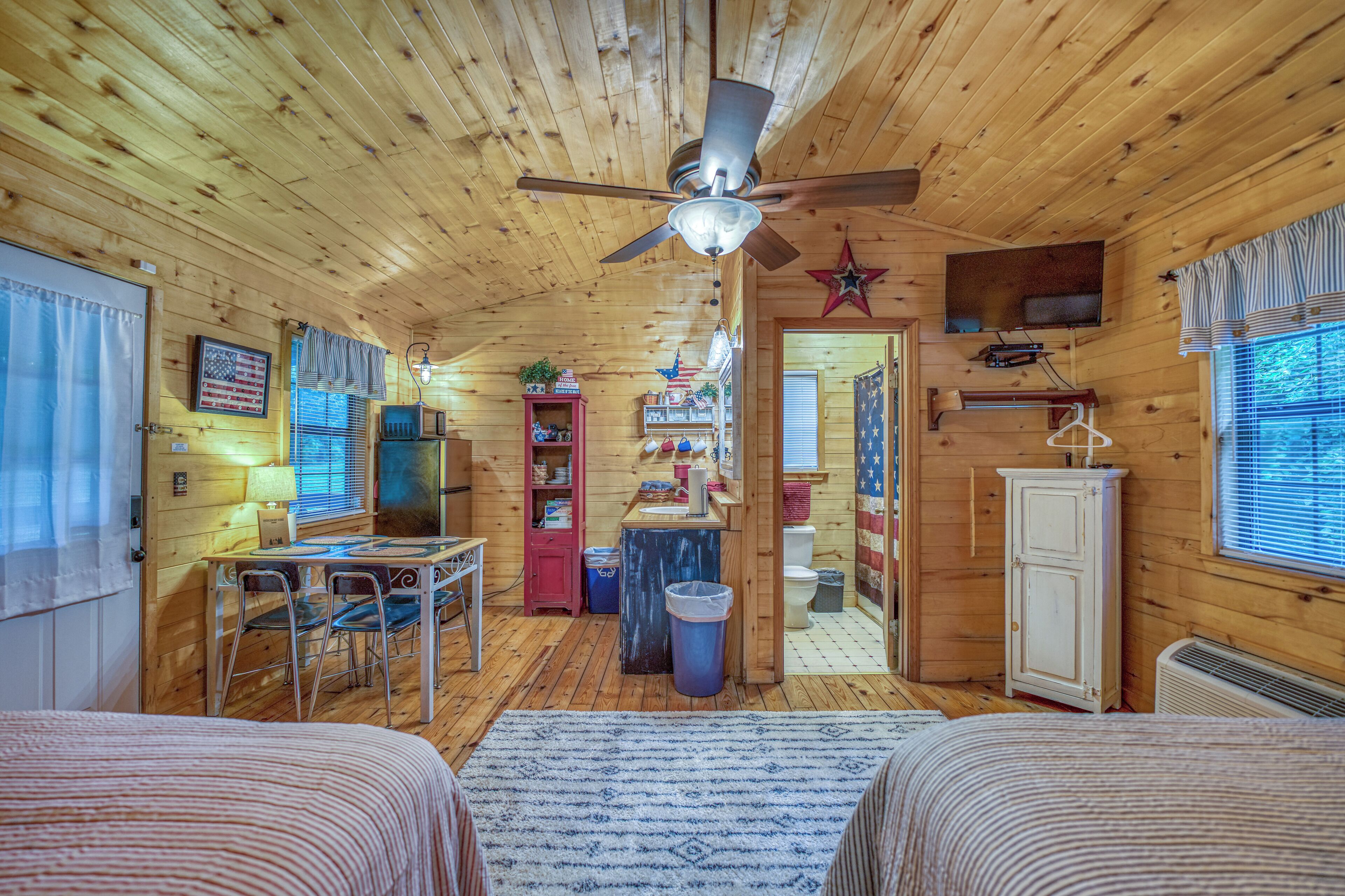 A cabin with a dining area, two beds, a ceiling fan, a TV, and a bathroom.