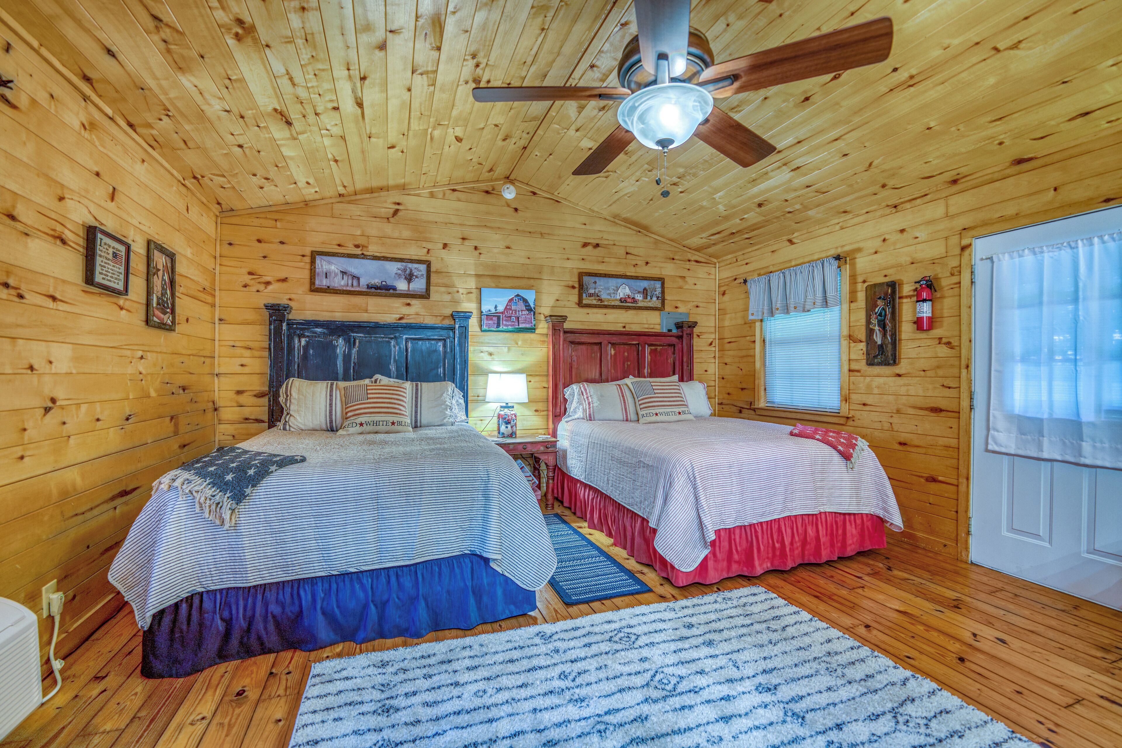 Une chambre de style cabane avec deux lits, des murs en bois, un ventilateur de plafond et une fenêtre avec des stores.