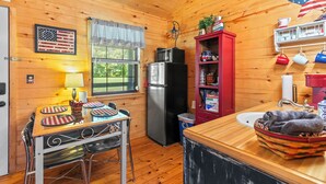 A wooden cabin interior with a dining table, chairs, a refrigerator, a microwave, a sink, and a shelf with mugs and a flag. - The Patriot Cabin close to the Buffalo National Park and River! Sleeps 4! (Harriet)
