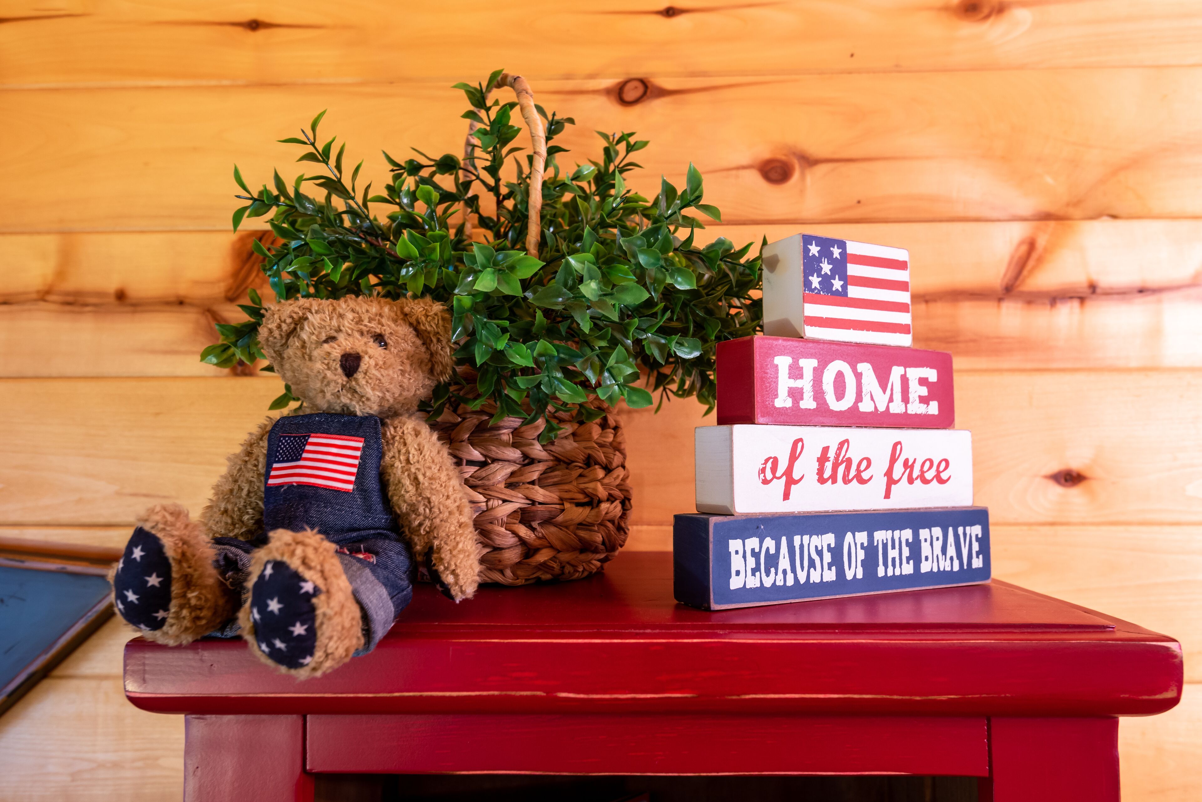A wooden bench with a teddy bear, a potted plant, and patriotic blocks spelling "HOME of the free BECAUSE OF THE BRAVE".