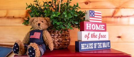 A wooden bench with a teddy bear, a potted plant, and patriotic blocks spelling "HOME of the free BECAUSE OF THE BRAVE".