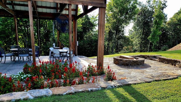A covered patio area with a stone fire pit, outdoor seating, and a garden with red flowers. - The Patriot Cabin close to the Buffalo National Park and River! Sleeps 4! (Harriet)