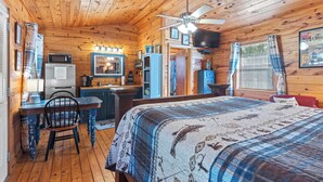 Een gezellig chaletinterieur met een houten vloer, een bed met een bedsprei met patroon, een eettafel met stoelen, een kitchenette en een plafondventilator.