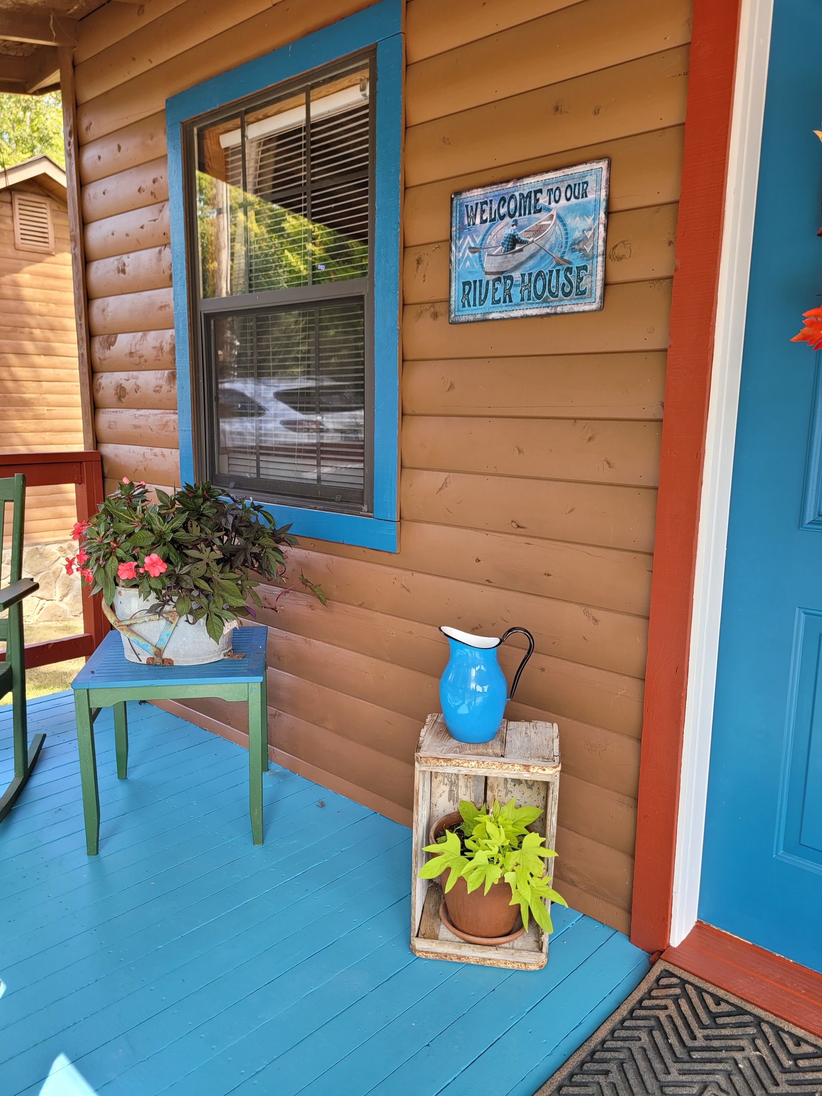 Una cabaña de madera con puerta y ventana azules, un letrero de bienvenida, una mesa azul con una planta en maceta y una jarra azul sobre un cajón de madera.