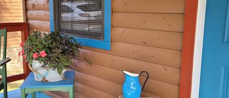 Una cabaña de madera con puerta y ventana azules, un letrero de bienvenida, una mesa azul con una planta en maceta y una jarra azul sobre un cajón de madera.