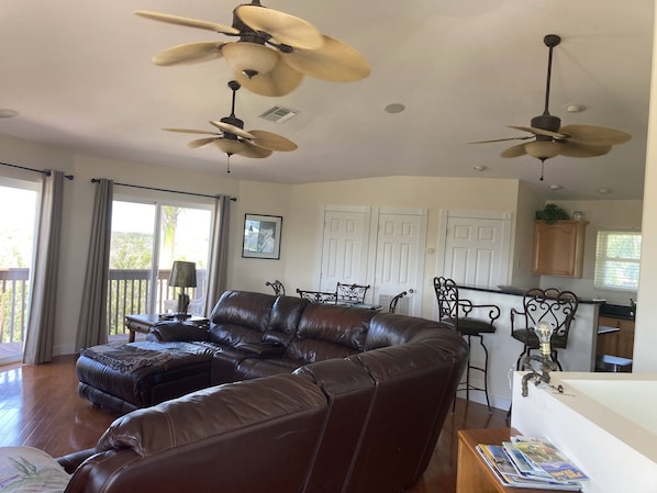 Ein geräumiges Wohnzimmer mit einer braunen Ledersofa, zwei Deckenventilatoren und einem Essbereich mit Tisch und Stühlen.
