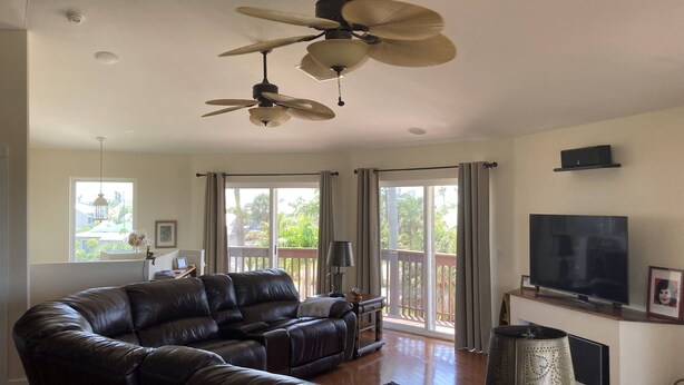 A spacious living room with a large leather sofa, a ceiling fan, a flat-screen TV, and a balcony with sliding glass doors.