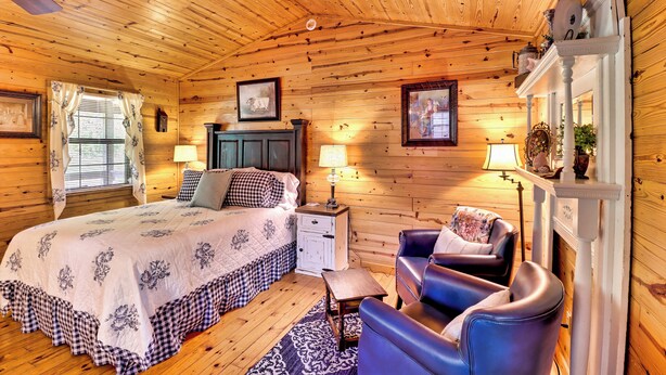 Un dormitorio de cabaña de madera con una cama, dos mesitas de noche, una silla, una chimenea y una ventana con cortinas.