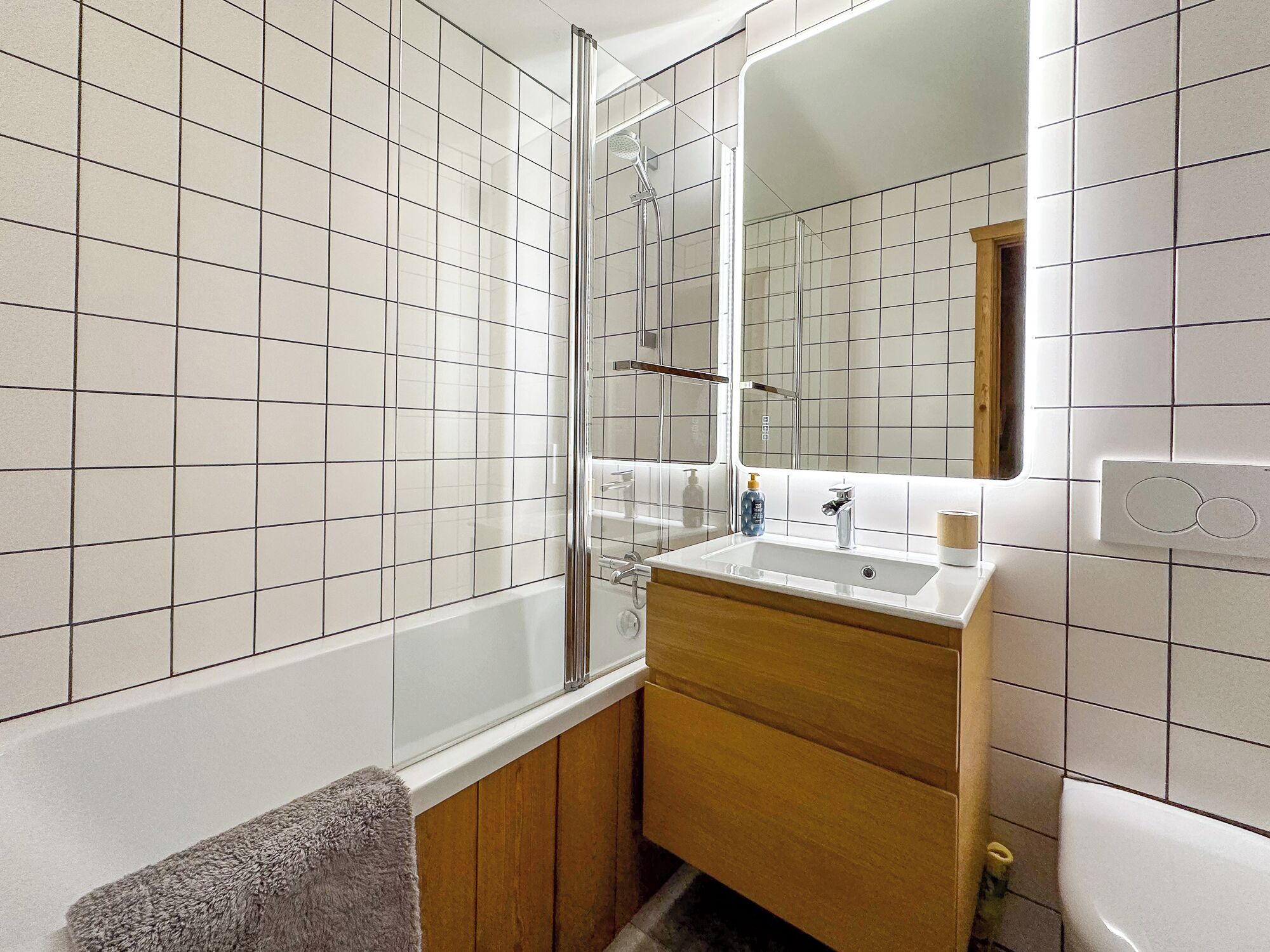 Une salle de bain avec des murs recouverts de carreaux blancs, un meuble-lavabo en bois, un lavabo, un miroir et une cabine de douche.