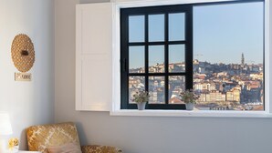 A room with a large window offering a city view, a patterned sofa, a small table with a lamp, and a decorative wall piece. - MyRiverPlace N.5 - PORTO Riverfront Apartment With Breathtaking Views (Vila Nova de Gaia)