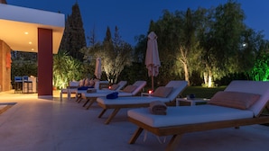 A poolside area with lounge chairs, umbrellas, and a building with a red pillar.