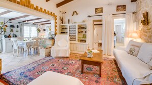Amplia sala de estar con un tapete grande y colorido, un sofá blanco, una mesa de centro de madera y un comedor con sillas y una mesa blancos.