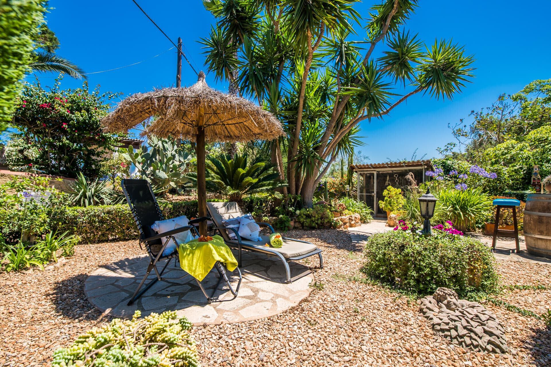 A patio with two lounge chairs, a thatched umbrella, and a small garden with various plants and a decorative statue.