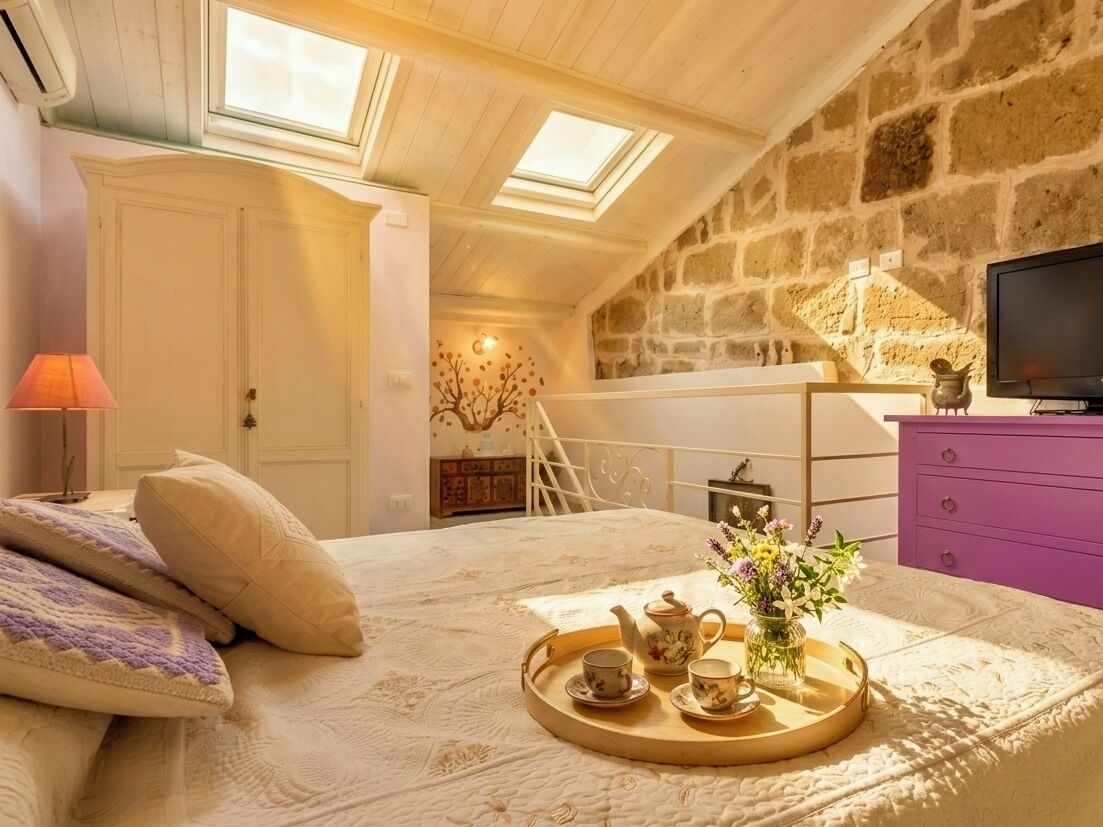 A cozy bedroom with a bed, a tray with tea and flowers, a TV, and a stone wall.
