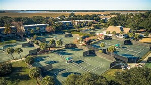 An aerial view of a residential area with multiple tennis courts, houses, and a beach in the background.