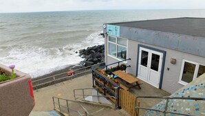 A coastal building with a staircase leading to a deck overlooking the sea, featuring a seating area with flowers and a view of the ocean. - The Plaice -  Wake up to panoramic sea views in The Plaice, Sheringham (Sheringham)