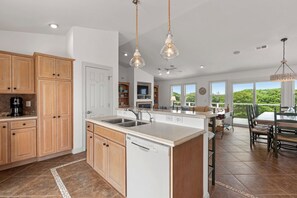 A spacious kitchen with wooden cabinets, a dishwasher, and a large island with a sink. There is a dining area with a table and chairs, and a view of greenery outside.