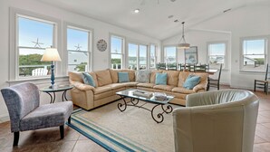 Ein geräumiges Wohnzimmer mit einer großen Eckcouch, einem Glastisch und einem Deckenventilator. Große Fenster bieten Blick auf Windkraftanlagen und eine weiße Außenwand.