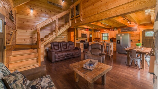 A spacious wooden cabin interior with a staircase, a living area with a sofa and chairs, a dining table, and a kitchen area. - Cozy Cabin, Great Location, Close to downtown, Private Hot Tub. (Gatlinburg)