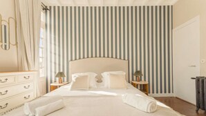 Een net opgemaakt bed met kussens en een dekbed, een dressoir met lades, en een gestreepte wand.