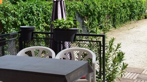 Un patio esterno con tavolo e sedie, una zona coperta e vista su siepe e alberi.