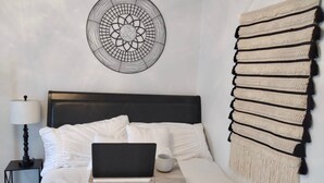 A neatly made bed with a laptop on it, a bedside table with a lamp, and a wall hanging. - The Charming Unit (5 minutes from Airport) (Carolina)