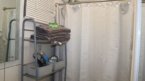 A bathroom with a shower curtain, a towel rack with folded towels, a shelf with baskets, and a mirror. - The Charming Unit (5 minutes from Airport) (Carolina)