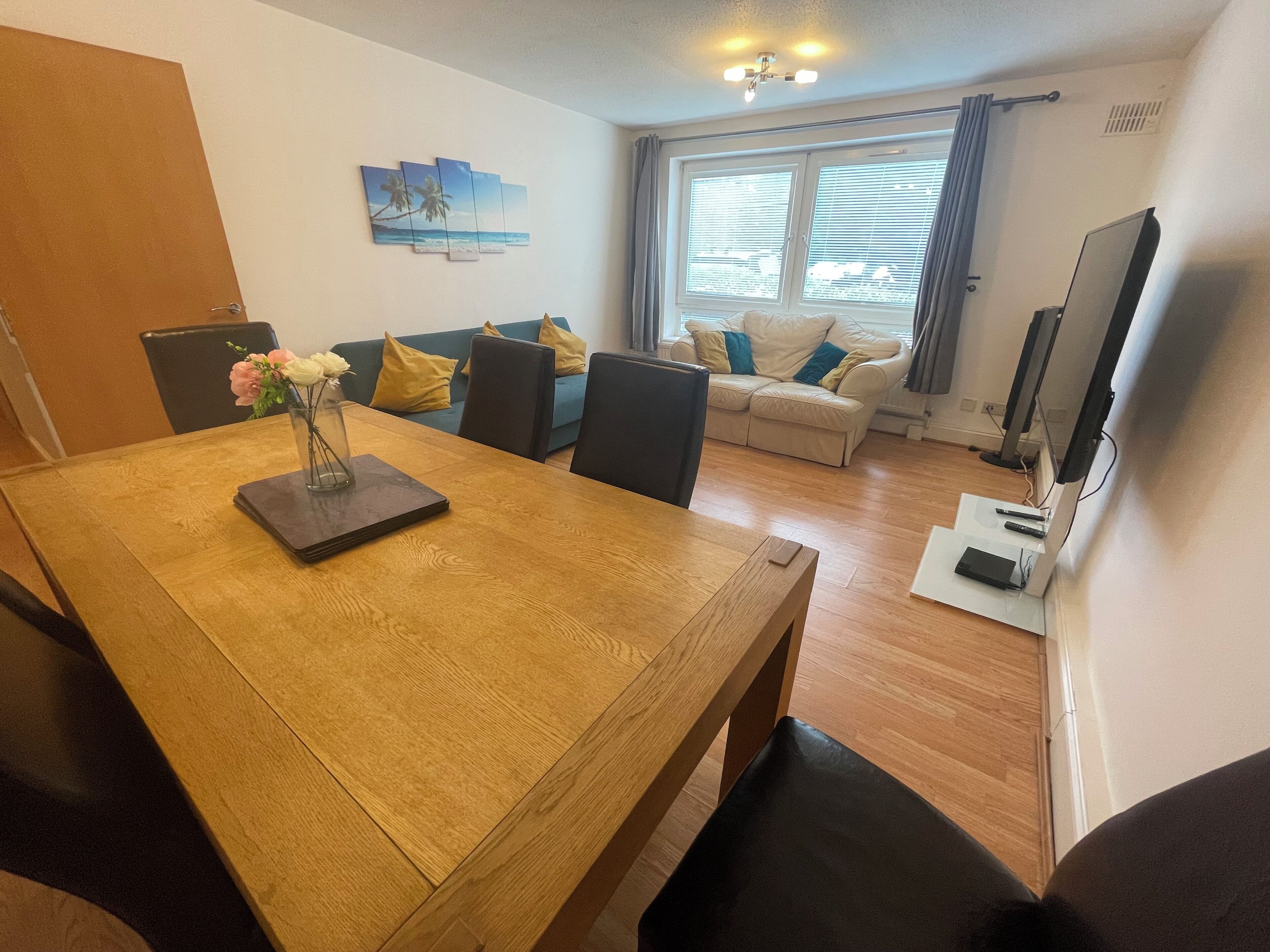 Un comedor con mesa de madera, sillas y un jarrón con flores. Hay un sofá con cojines y una televisión al fondo.